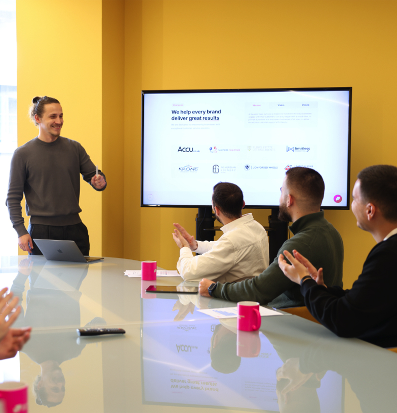 A presenter with a confident smile leads an interactive session in front of a screen displaying client success stories, with engaged team members applauding in a meeting room.