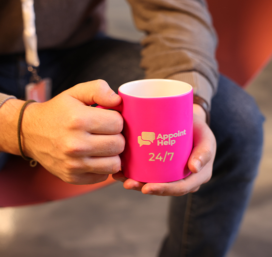 Hands holding a bright pink AppointHelp mug with the text '24/7', symbolizing round-the-clock customer support availability.