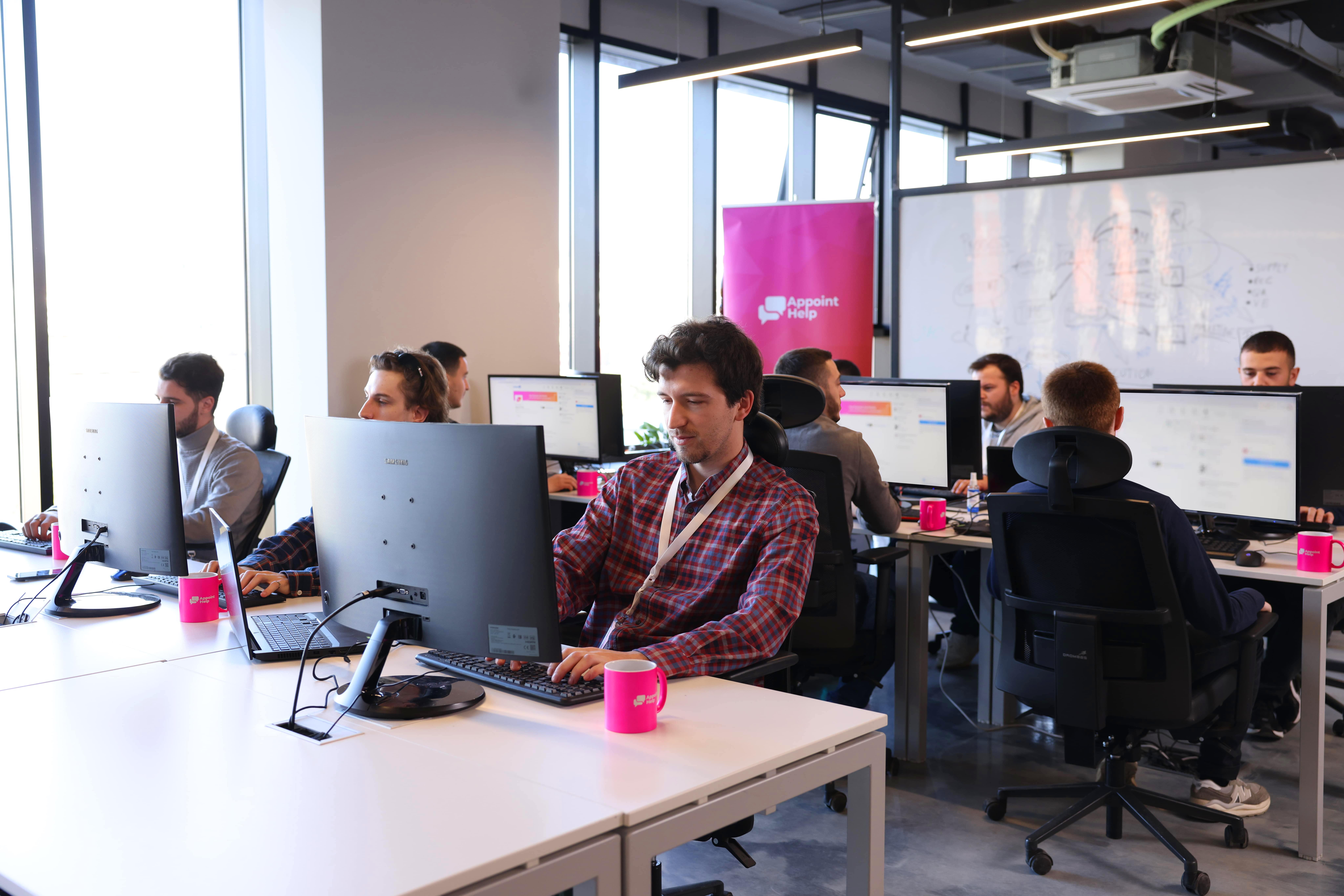 Team of customer service representatives providing outsourced live chat support in a modern office setting with branded AppointHelp mugs on desks.