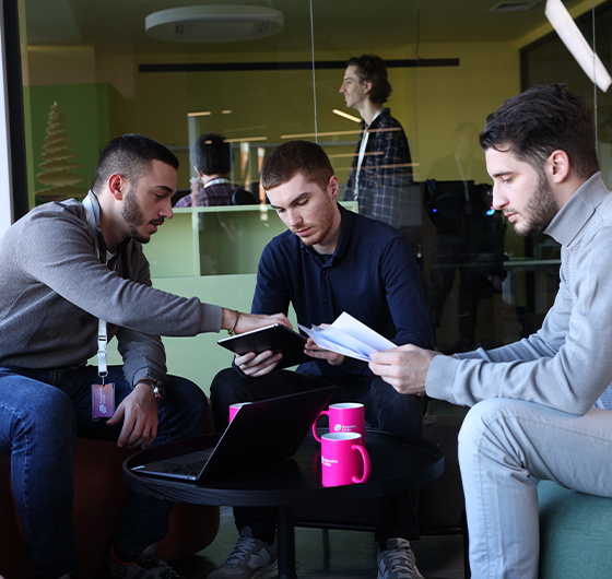 Three team members engaged in a collaborative discussion over documents and digital devices, with a branded AppointHelp mug on the table, in a modern office setting.