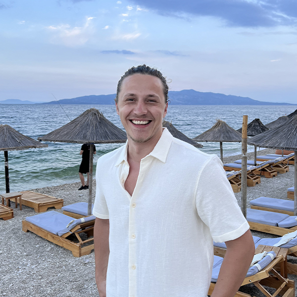 A smiling man in a casual white shirt standing on a beach with thatched umbrellas and calm sea in the background, exuding a relaxed and approachable demeanor.