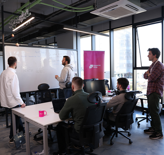 A team meeting in progress in a contemporary office space, with one member presenting at the whiteboard and others engaged in discussion, featuring the AppointHelp brand.