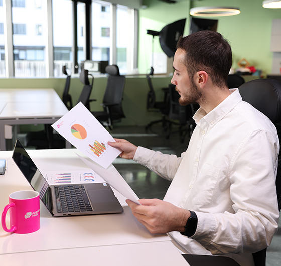 Focused professional analyzing statistical reports beside a laptop and a bright pink AppointHelp mug on a white office desk.