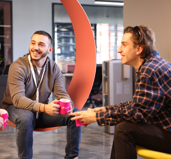 Two cheerful male colleagues enjoying a break with branded AppointHelp mugs, in a casual office lounge area with a distinctive red sculpture.